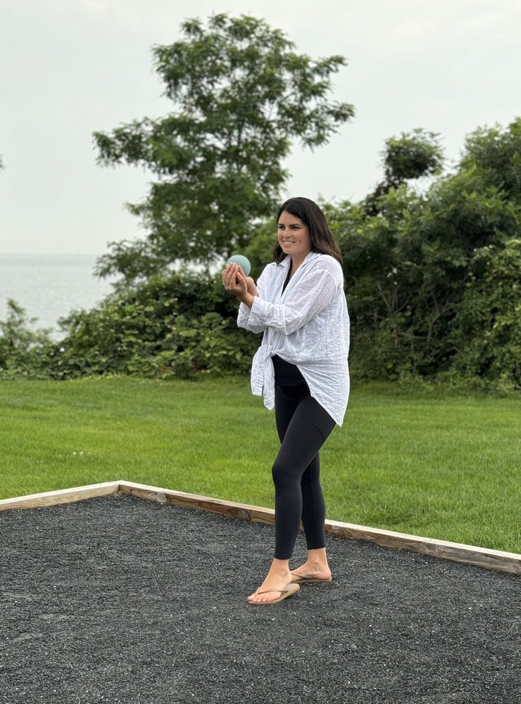 woman holding a bocce ball wearing black leggings and a white shirt.