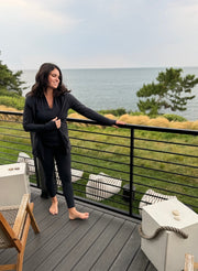 woman on a deck with water in the background. she is wearing a black outfit with leggings, a cardigan and a black top