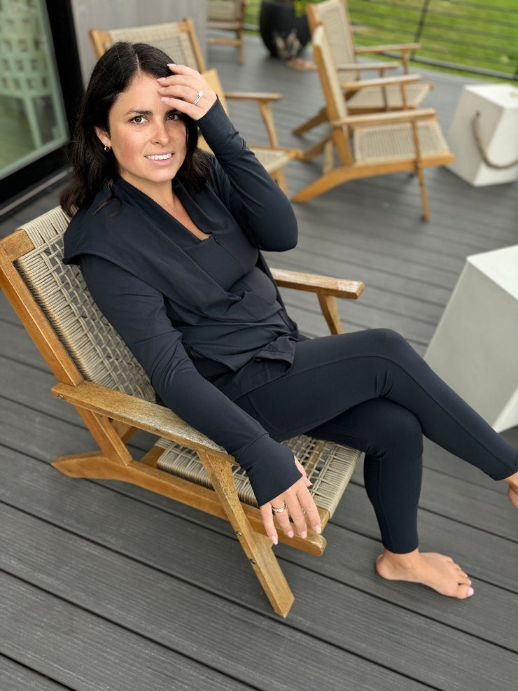 woman sitting on a deck chair wearing black leggings with stitching and pocket details, an a black open cardigan and activewear top