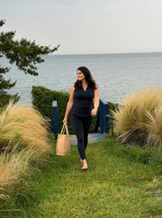 woman walking toward us with water i the background. she carries a tote and is wearing black workout leggings and a top.