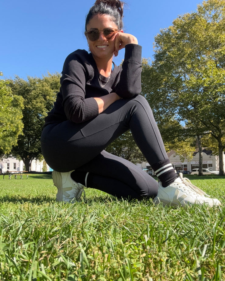 Woman in athletic wear and meira active smooth duo leggings with pockets  stretching outdoors on grass with trees and blue sky in the background
