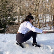 Woman turned to the side and crouching down wearing full length black leggings. Scenery shows trees and snow.