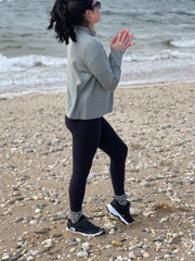 Woman standing on shoreline in the sand with ocean in the background wearing a sweater, black full length leggings and sneakers. She is turned to the side.
