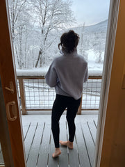 Woman facing away wearing ankle length black leggings and a blue sweater. She is standing on a deck looking out towards snow covered trees and mountains.